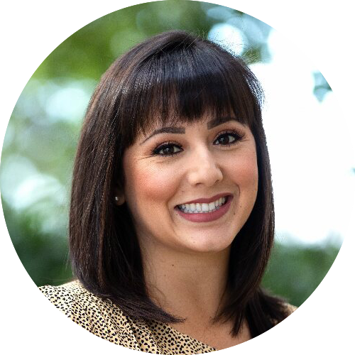 A smiling woman with dark hair and bangs, wearing a patterned top, against a blurred green background.