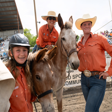 Three women in cowboy hats pose with horses at a rodeo event, showcasing a spirit of camaraderie and equestrian culture.