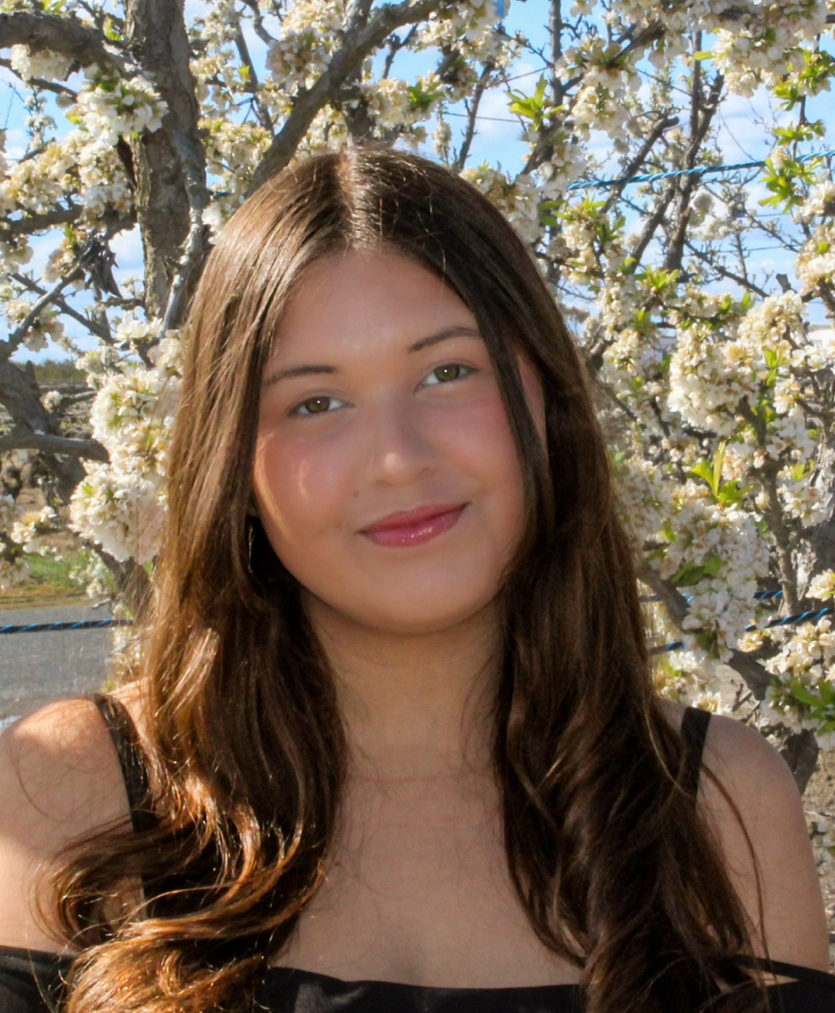 A young woman with long, wavy hair smiles softly in front of a backdrop of blooming flowers.