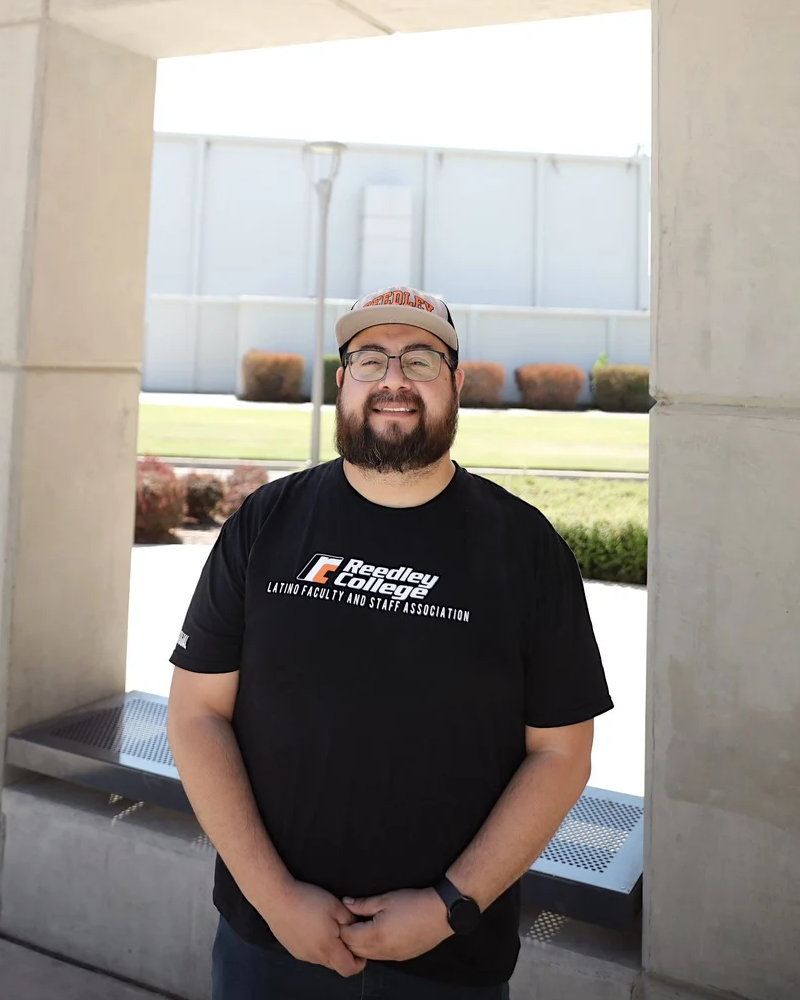 A man wearing a black t-shirt with the logo of the Reedley College Latino Faculty and Staff Association, standing outdoors in front of a minimalist concrete structure.