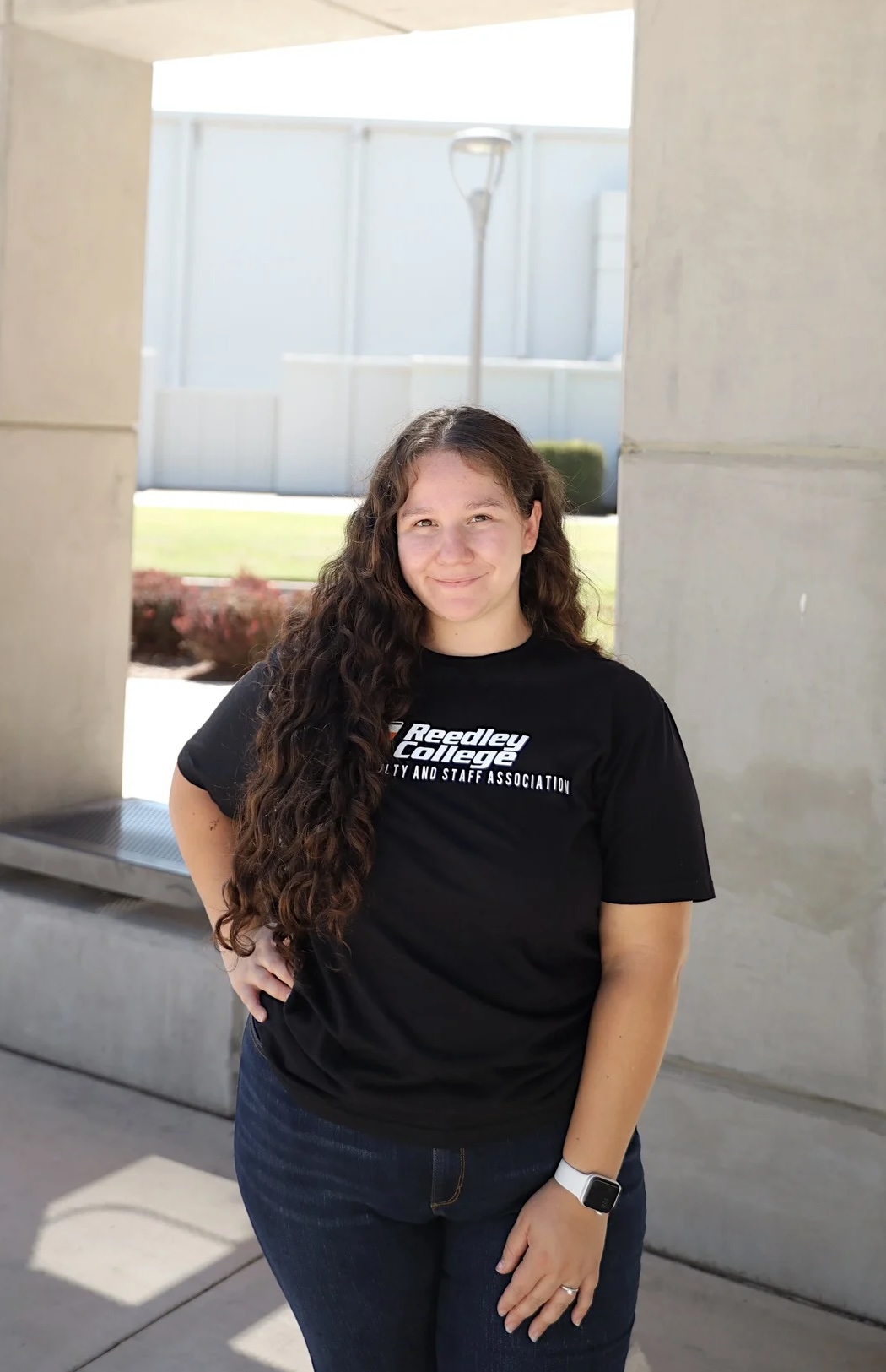 A man wearing a black t-shirt with the logo of the Reedley College Latino Faculty and Staff Association, standing outdoors in front of a minimalist concrete structure.