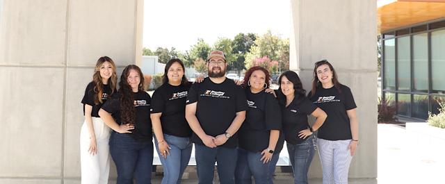 Team photo featuring eight diverse individuals in matching black t-shirts, posing together outdoors in a modern setting.