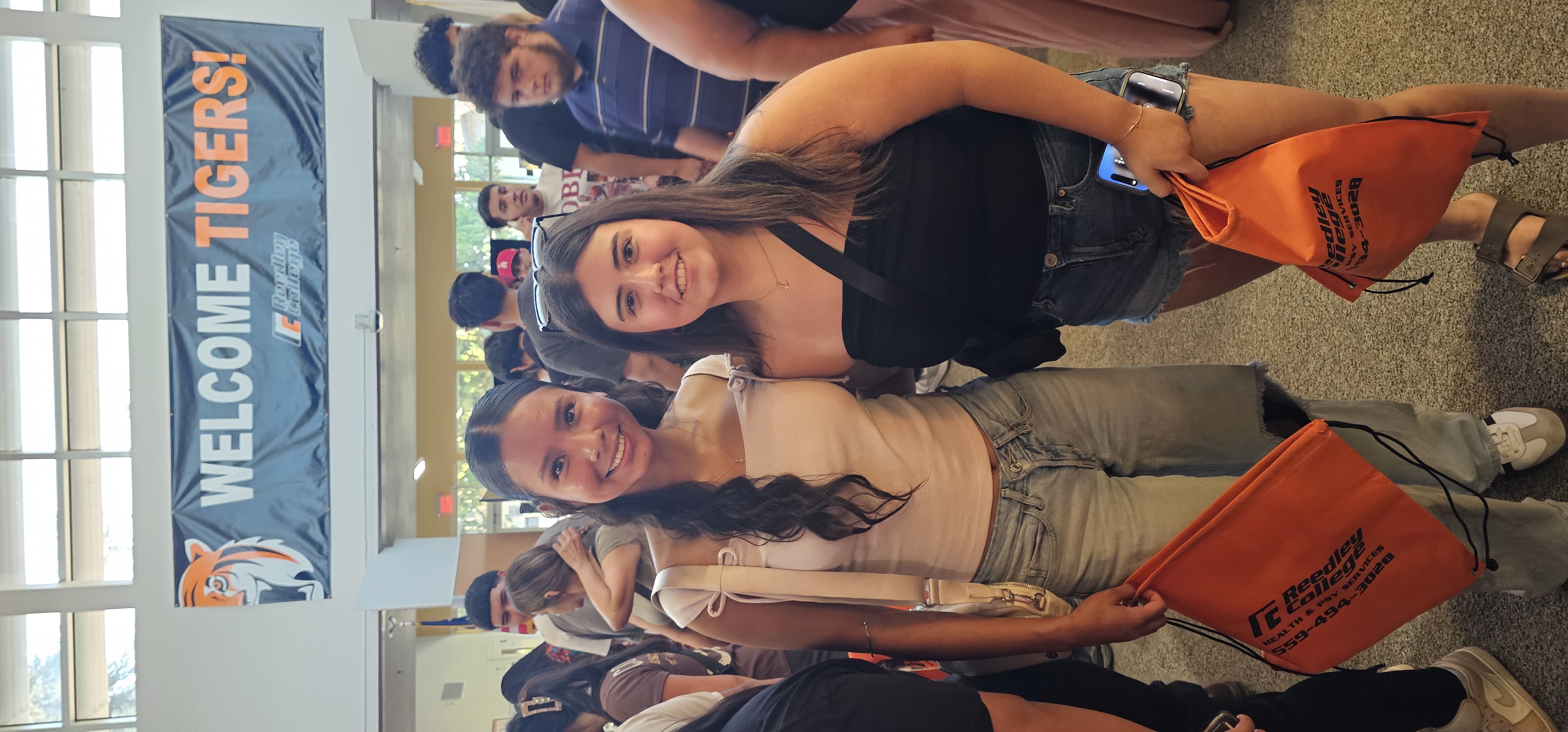 Two female students posing together at a campus welcome event with a "Welcome Tigers" banner in the background. Each student is holding an orange promotional bag.