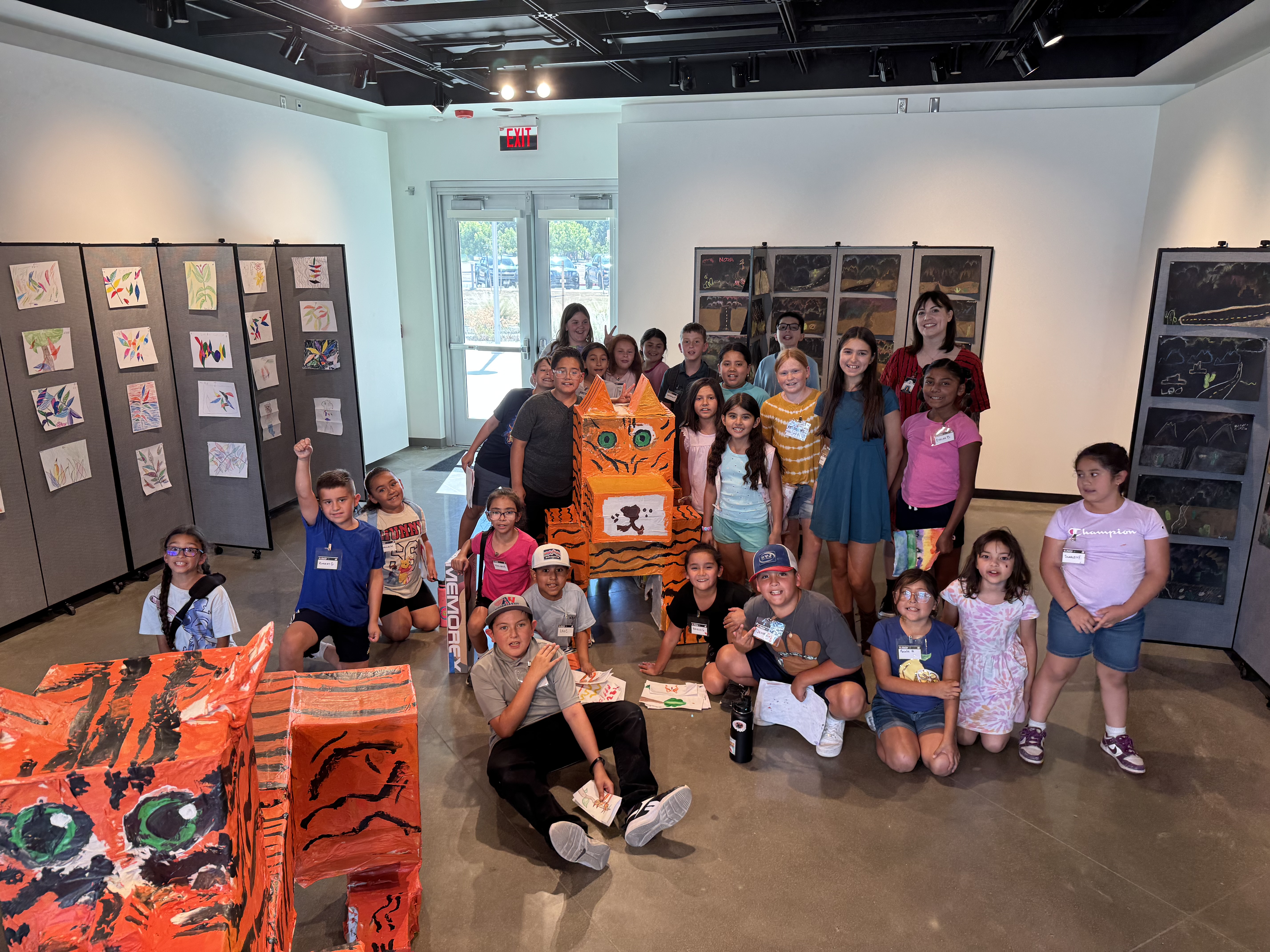 A group of children and adults pose in an art gallery surrounded by colorful artwork, featuring a large orange tiger sculpture in the foreground.